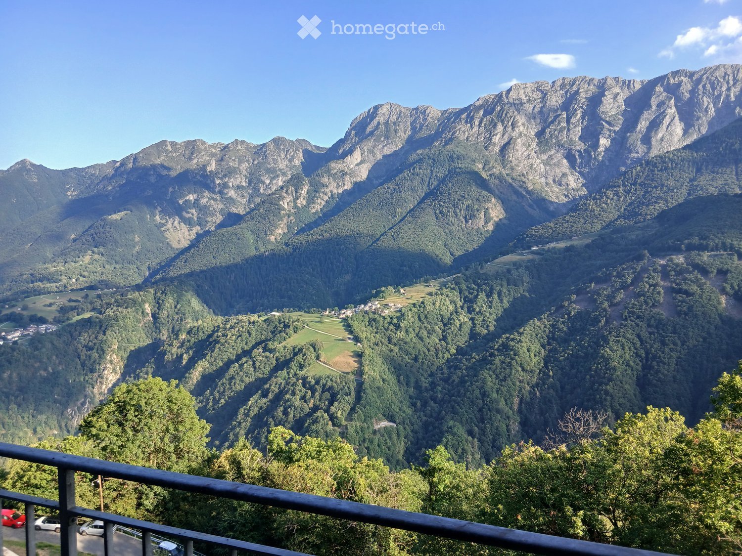 Mountain range, lush greenery, blue sky, black fence, parked cars