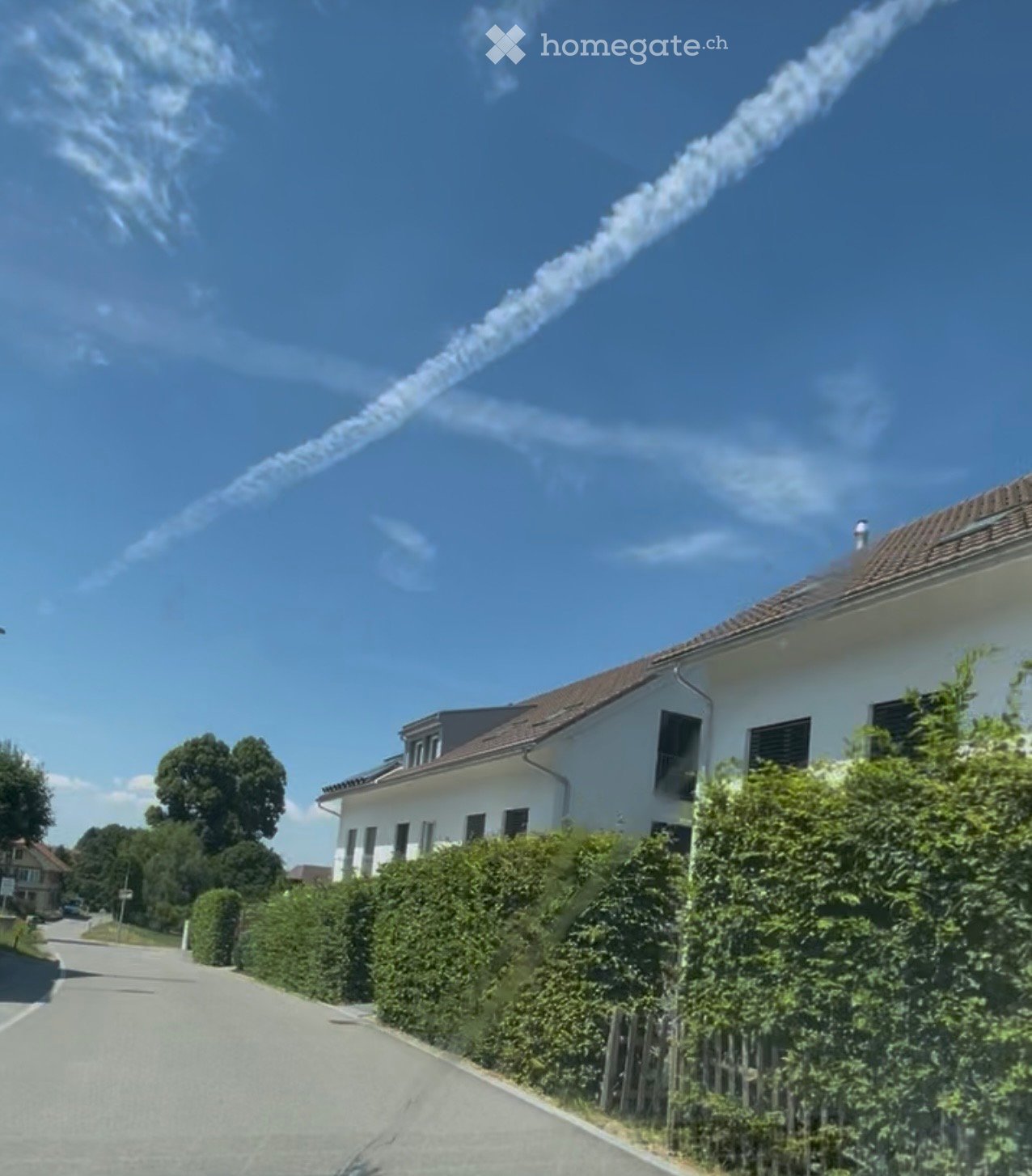A two-story white house with a brown roof, surrounded by greenery and a clear blue sky with contrails from an aircraft.
