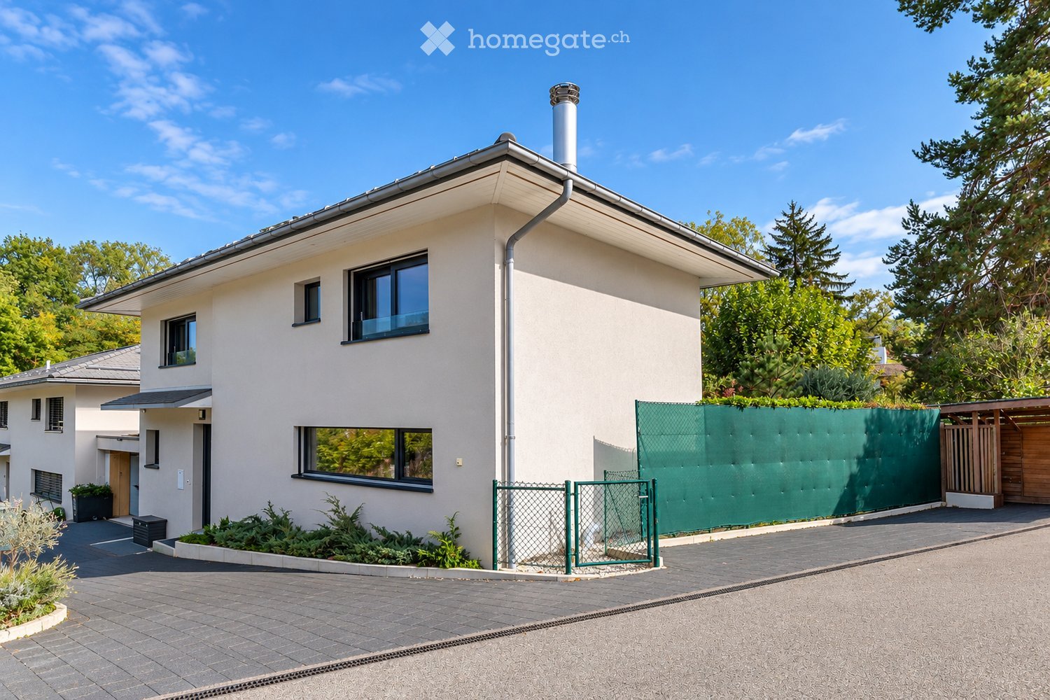 Modern two-story house with white exterior, large windows, and a green fence surrounding the property.