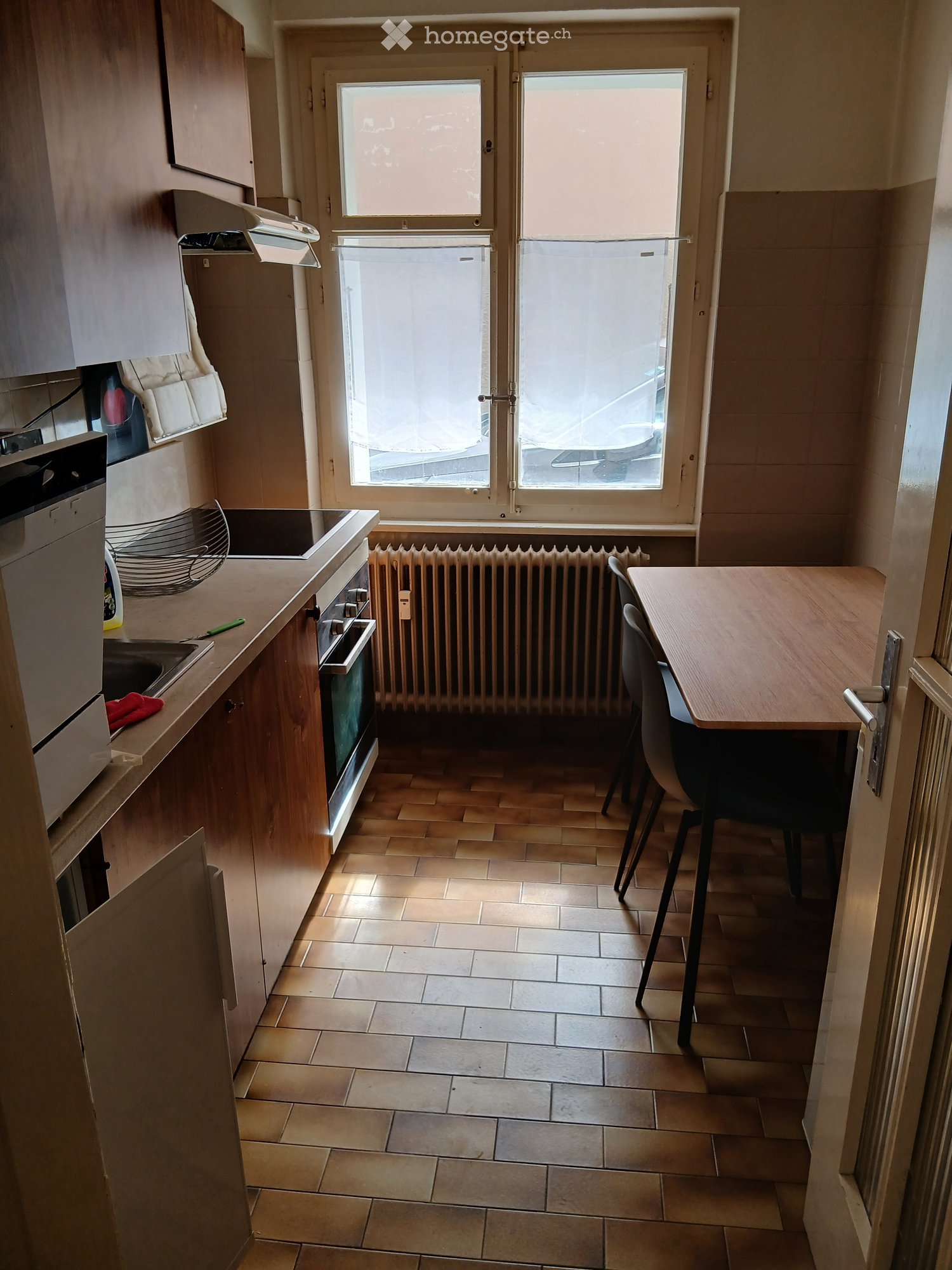 Modern kitchen with tiled floor, wooden cabinets, sink, oven, dishwasher, small dining table with chairs, and a window with blinds.