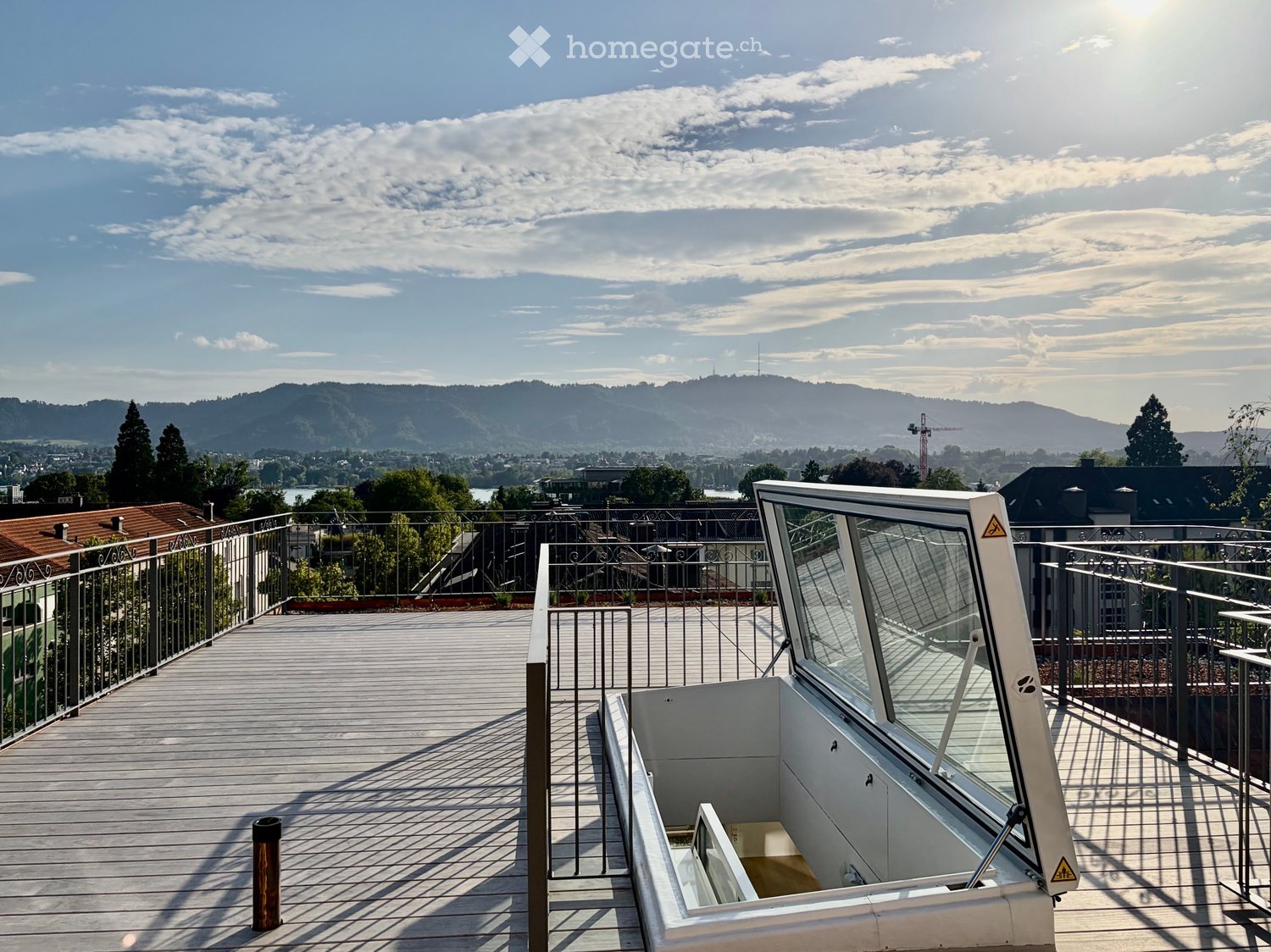 A modern rooftop terrace with wooden decking, metal railings, and a scenic view of a city and mountains in the background.