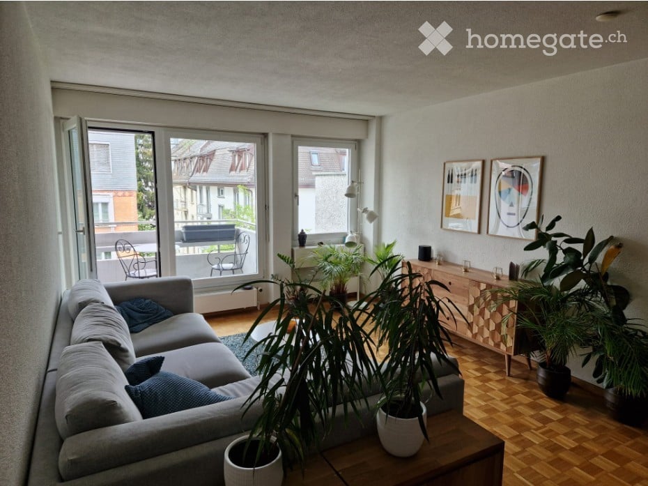 Modern living room with a grey sectional sofa, coffee table, side table, potted plants, and a balcony with outdoor seating.
