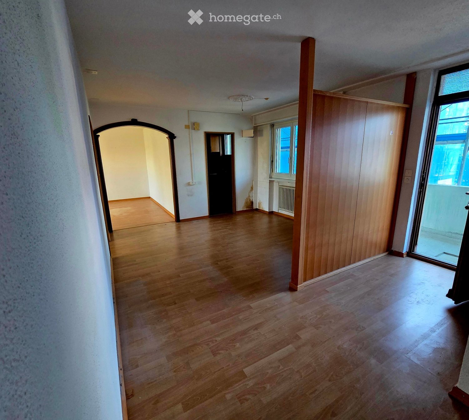 Empty hallway with wooden flooring, white walls, and a large window at the end. There are doors leading to other rooms and a visible radiator on one wall.