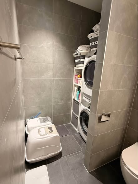 Modern bathroom with tiled walls and floor, featuring a toilet, washing machine, laundry basket, and pet litter box.