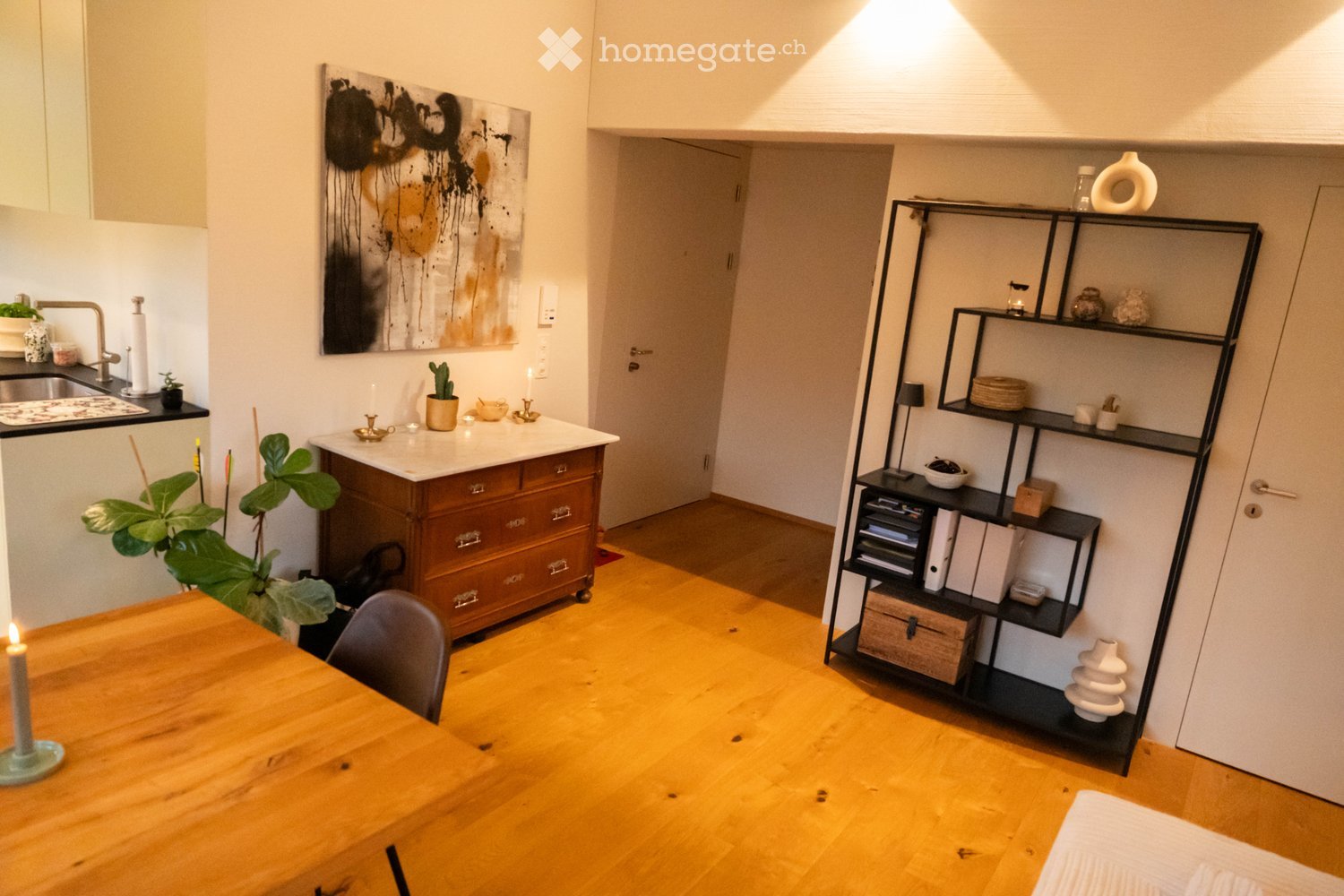 A modern, open-plan living and dining area with wooden flooring, a large wooden dining table, a black shelving unit, a sink, and a decorative art piece on the wall.