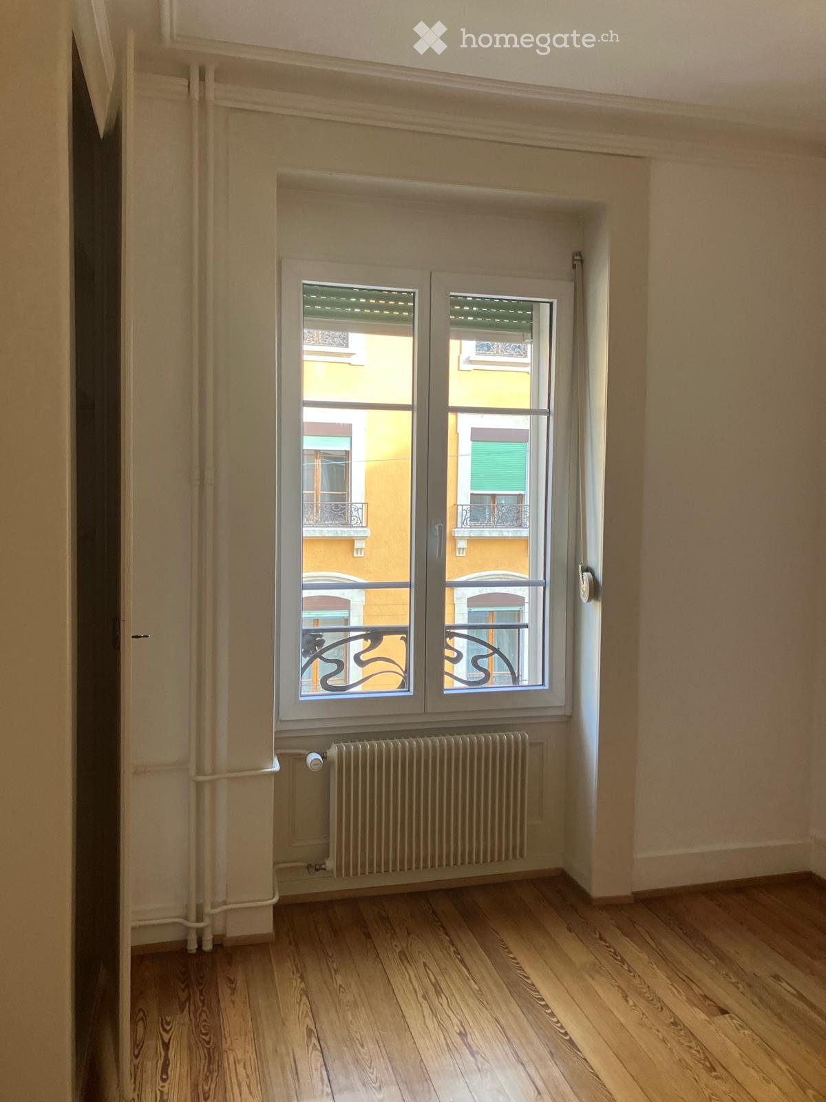 Empty room with wooden flooring, large window with double doors leading to a balcony, radiator below the window, and light-colored walls.
