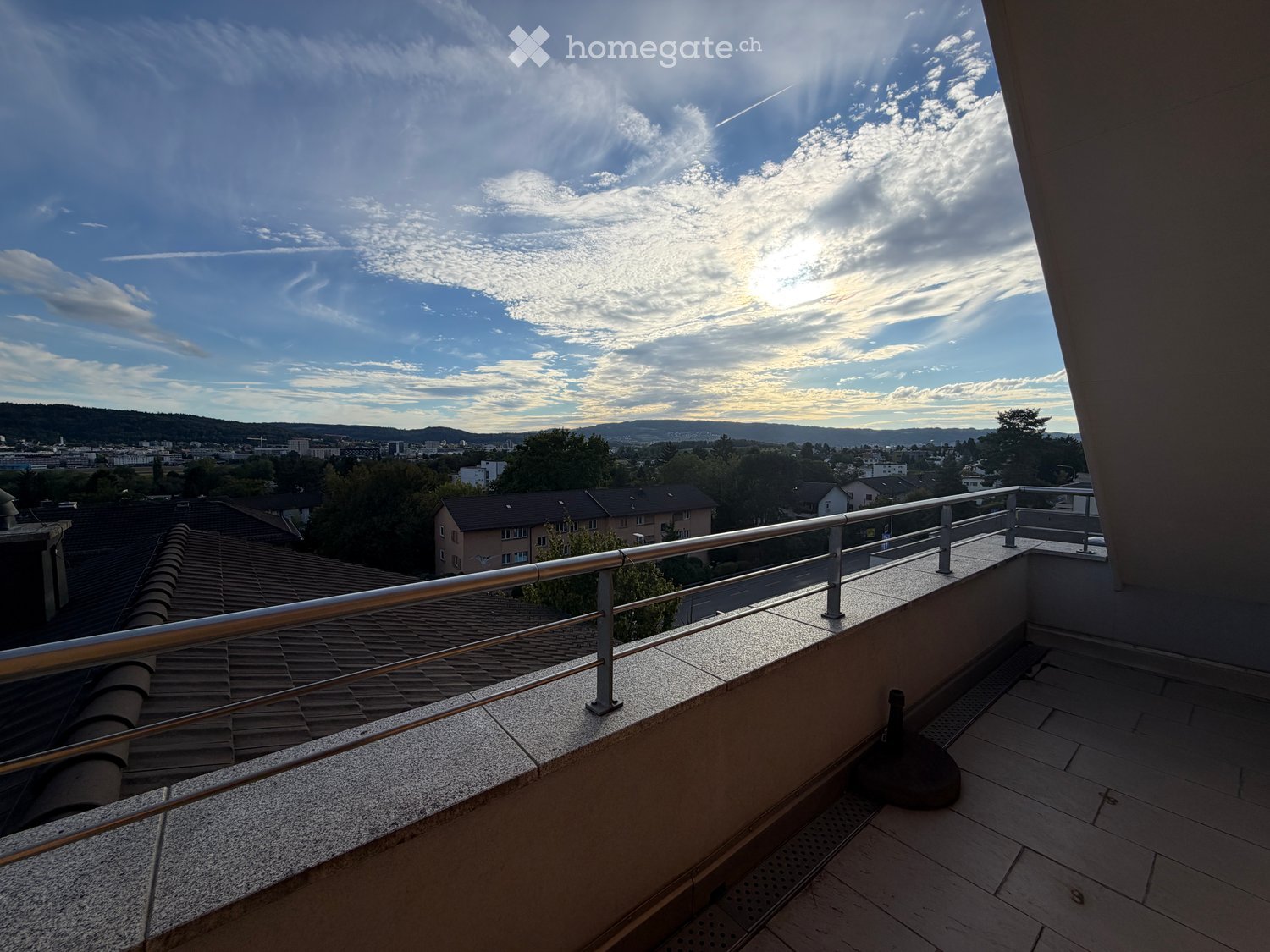 Balcony with metal railing, tiled floor, and a view of the cityscape and surrounding hills under a partly cloudy sky.
