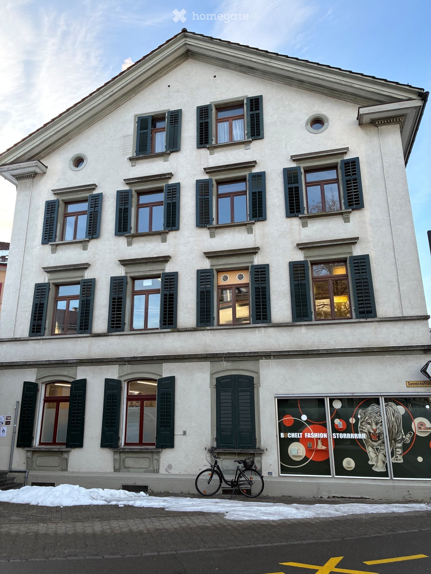 Three-story building with white facade, multiple windows with green shutters, and a storefront with a large tiger graphic on the window display. A bicycle is parked in front of the building.