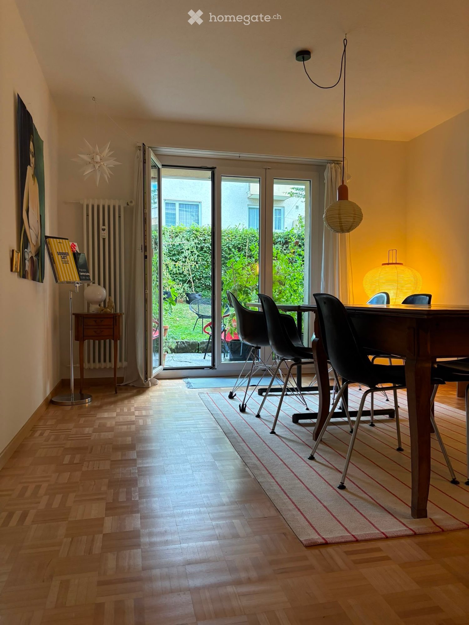 Bright dining area with wooden flooring, large sliding glass doors opening to a garden, a wooden dining table with black chairs, a hanging lamp, a floor lamp, a radiator, a globe, and a painting on the wall.