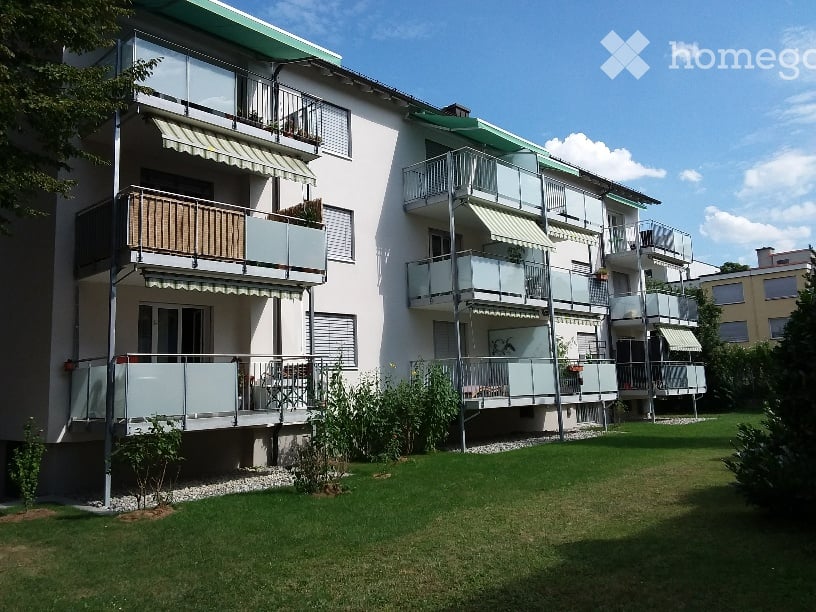 Modern multi-story apartment building with balconies, green roof, and surrounding lawn.