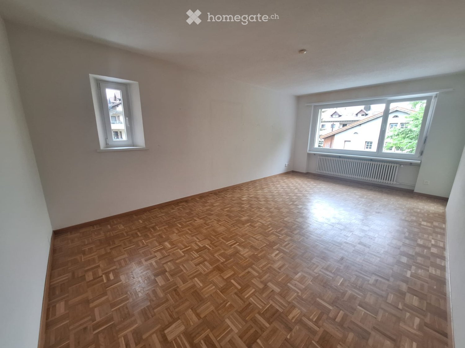 Empty room with wooden herringbone flooring, light-colored walls, and two windows offering a view of neighboring buildings.