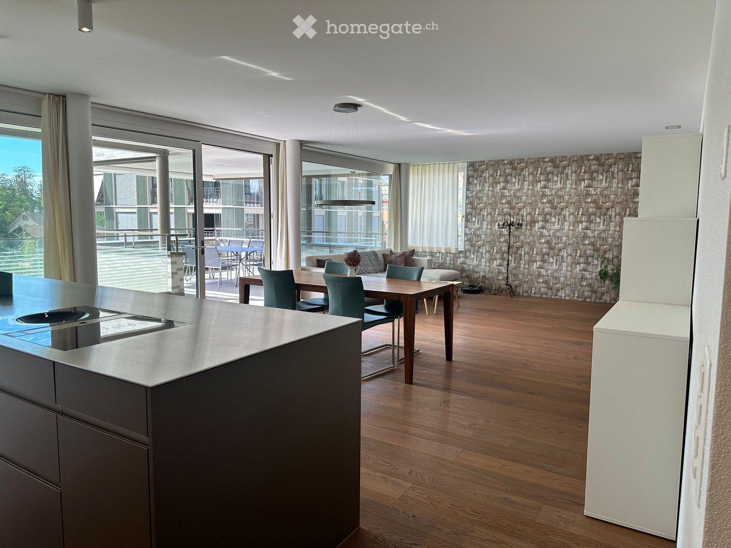 Modern open-plan living and dining area with a kitchen island, dining table, sofa, and ottoman, connected to a balcony with outdoor seating.