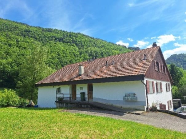 Single-story house with a red-tiled roof, white walls, and a small porch. Surrounded by green grass and trees with a mountain backdrop.