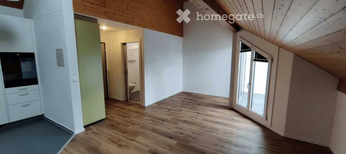 Empty room with wooden flooring, white walls, and a sliding glass door leading outside. Adjacent areas include a kitchen and a bathroom.