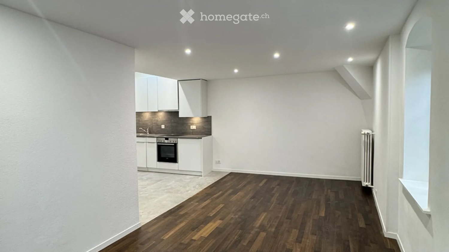 Modern, minimalist room with wooden flooring, white walls, and a compact kitchen area featuring cabinets, an oven, and a faucet.