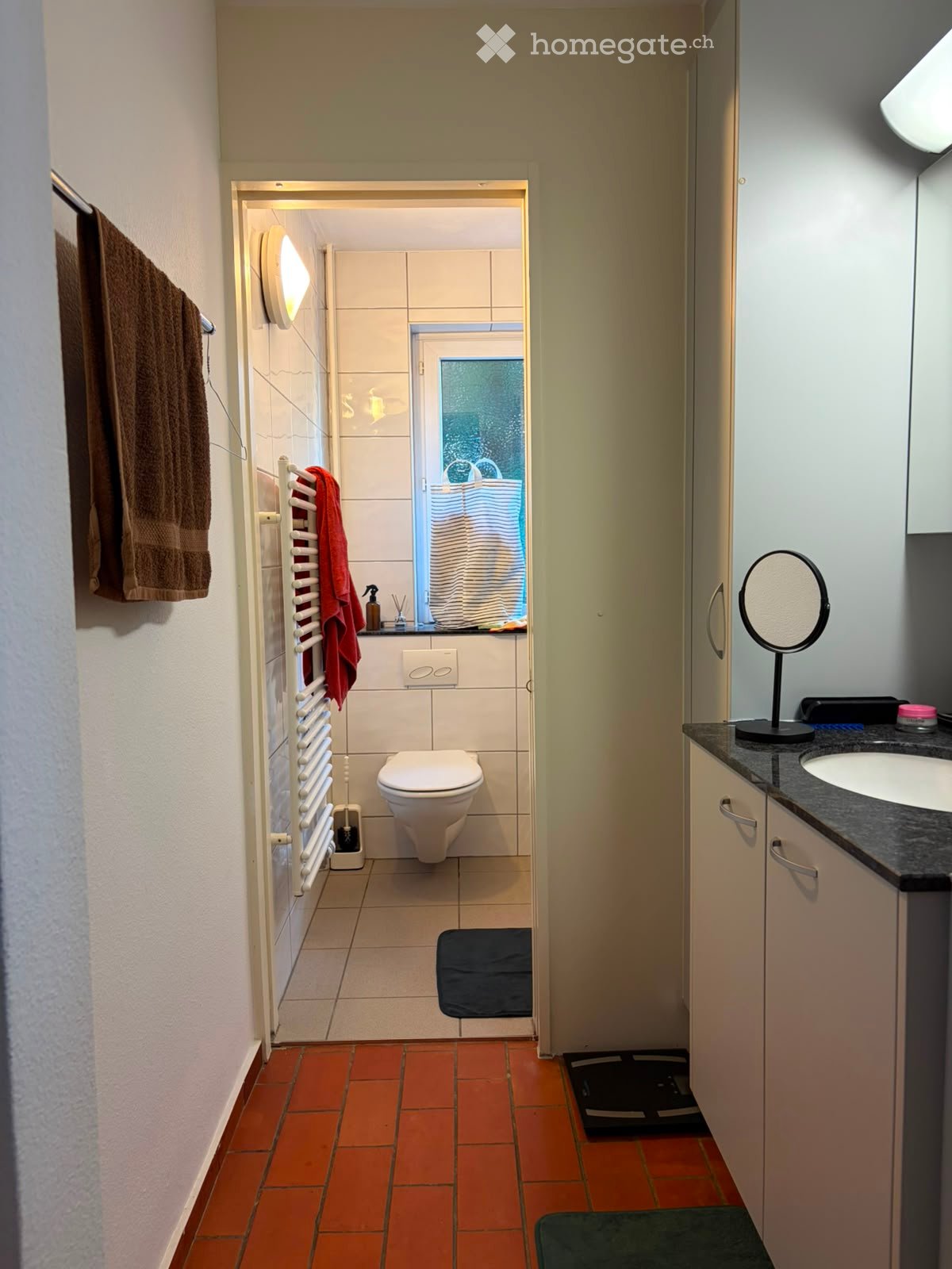 Modern bathroom with a toilet, sink, mirror, and towel. Features a radiator, tiled walls, and a window with a frosted glass design.