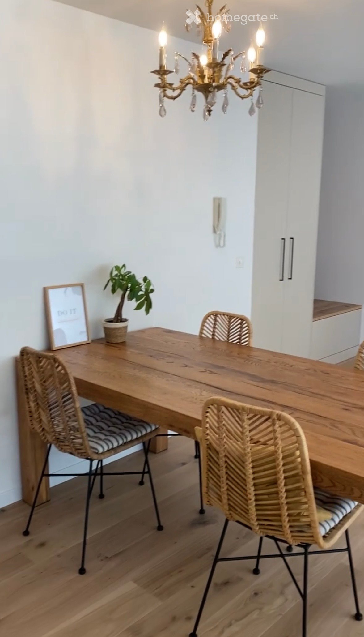 Dining area with a wooden table, four wicker chairs, a potted plant, a picture frame, and a chandelier.