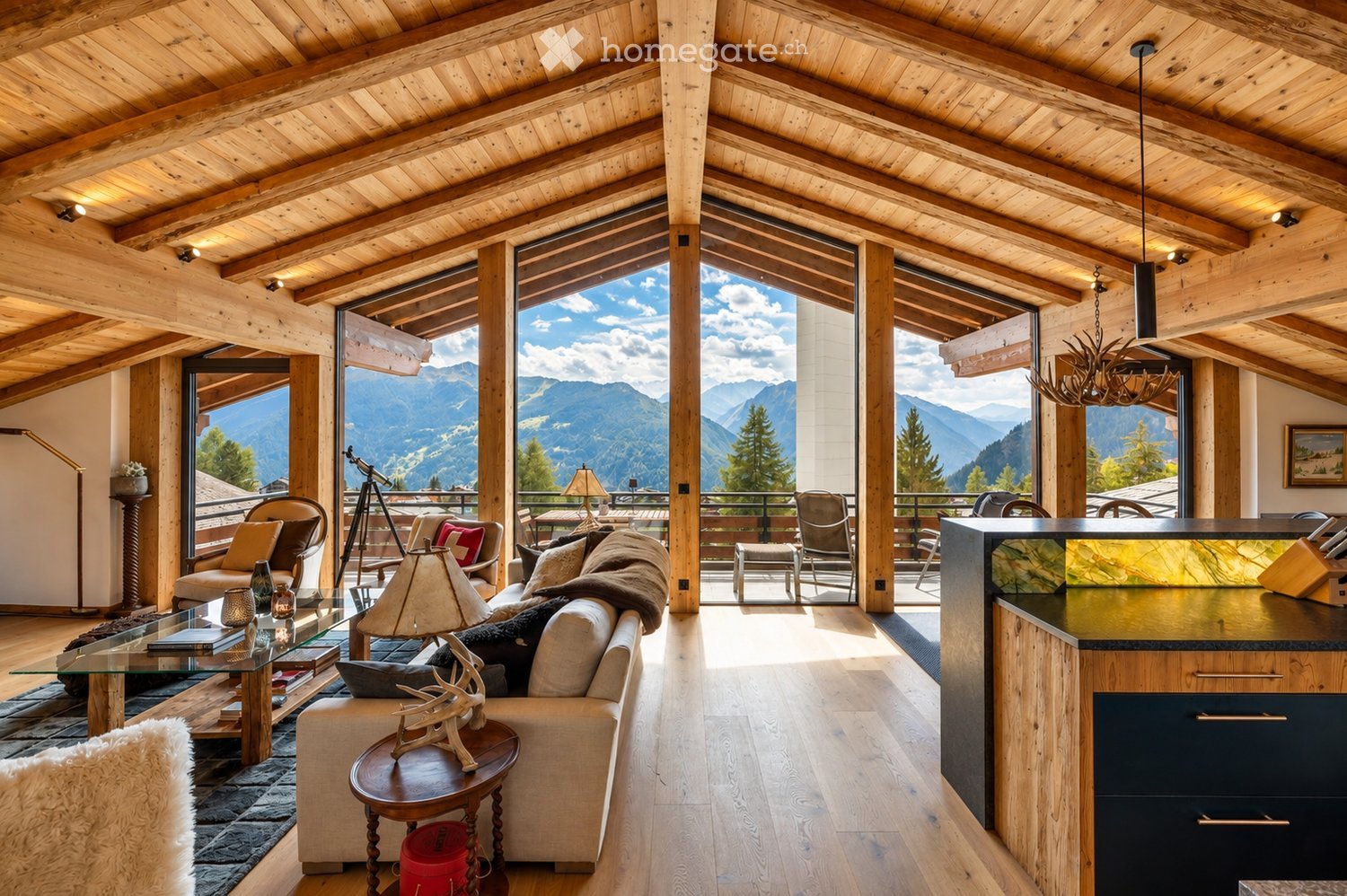 A spacious, well-lit living room with exposed wooden beams and large windows offering a scenic mountain view. The room features a cream-colored sofa, a glass coffee table, a wooden side table with a decorative deer head, and a modern kitchen island. Addit