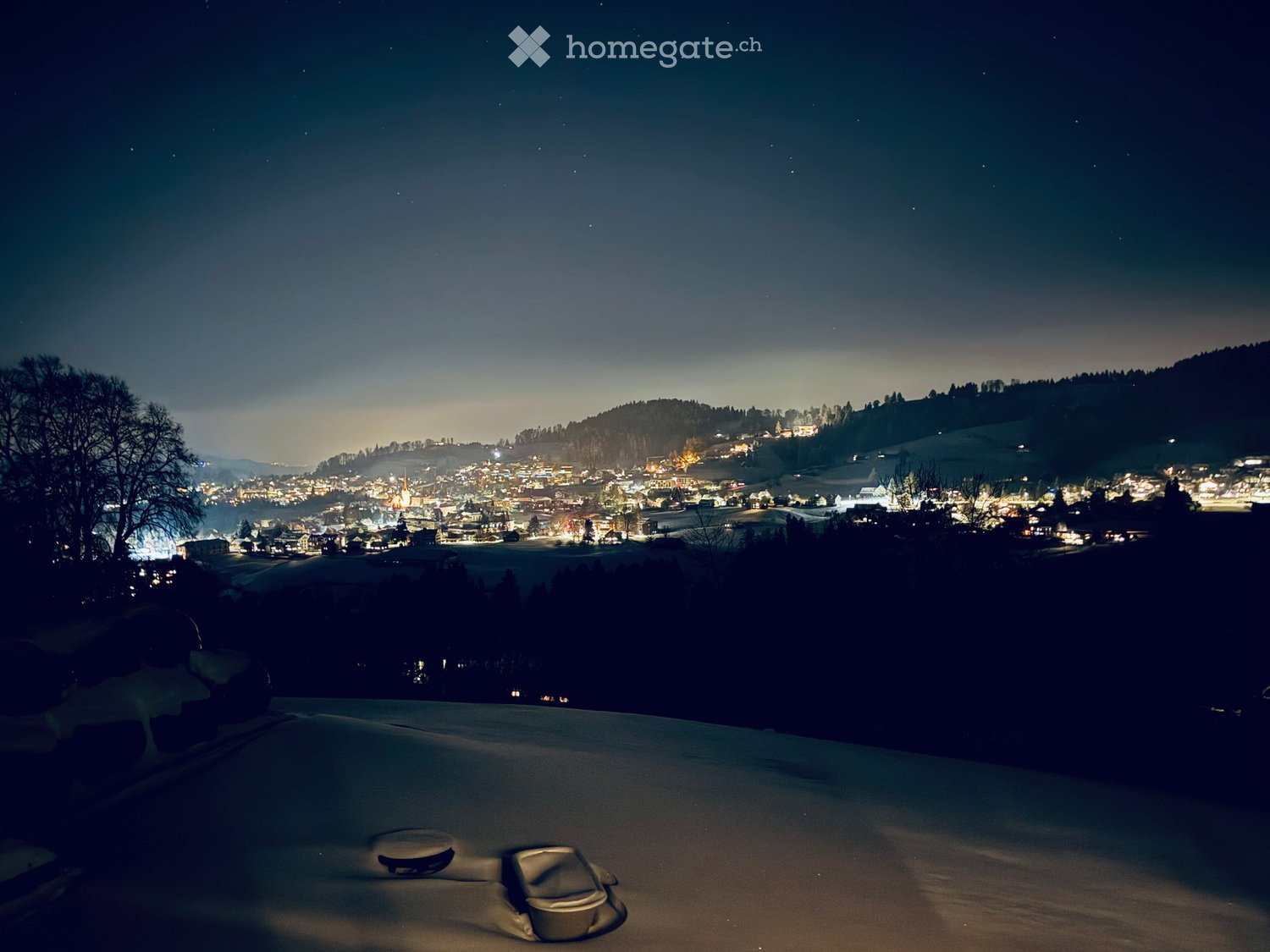 Nighttime view from a snow-covered balcony or terrace overlooking a town with illuminated buildings and surrounding hills.