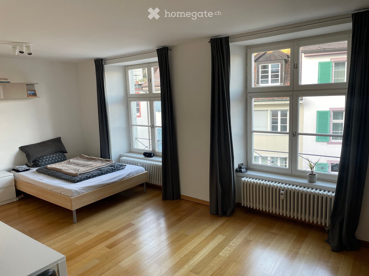 Cozy bedroom with wooden flooring, large windows with curtains, a single bed with pillows and blankets, a small shelf, and a potted plant on the windowsill.