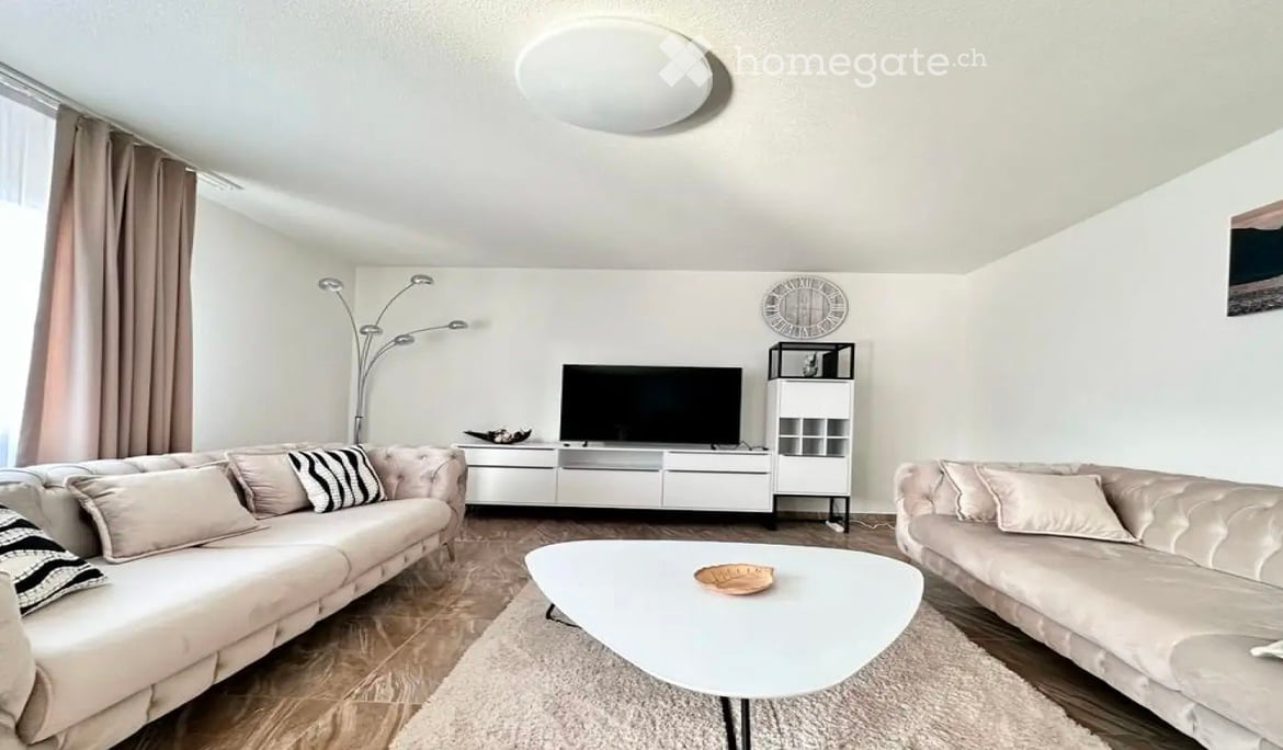 Modern living room with light-colored walls, wooden flooring, and large windows. Features a cream-colored sofa, a white coffee table, a television, a floor lamp, and a small shelf.