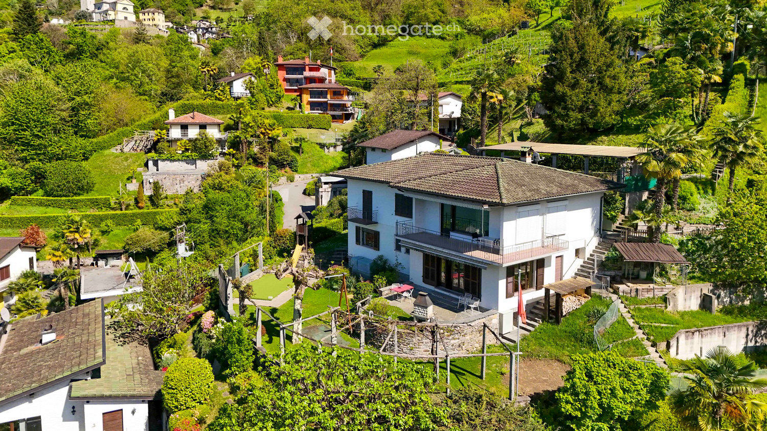 Aerial view of a residential area with multiple houses surrounded by lush greenery and trees. The houses have varied architectural styles and are situated on a hilly terrain.