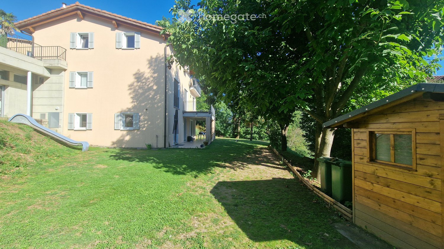 A well-maintained garden area with a lawn, a wooden shed, and a two-story house with balconies and shutters in the background.