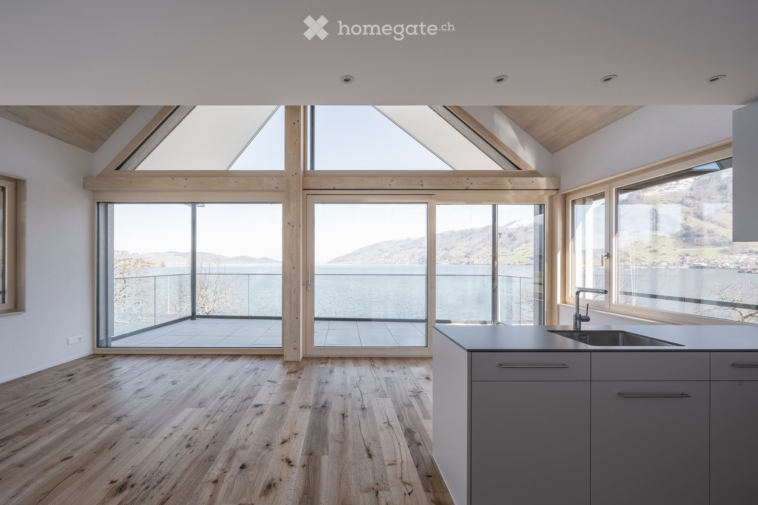 Modern kitchen with wooden flooring, grey cabinets, and large windows offering a scenic view of a lake and mountains.