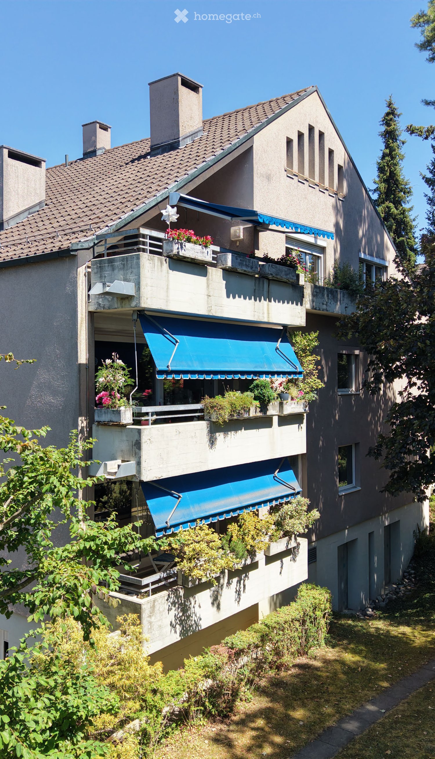 A multi-story residential building with balconies, blue awnings, and flower boxes, surrounded by greenery.