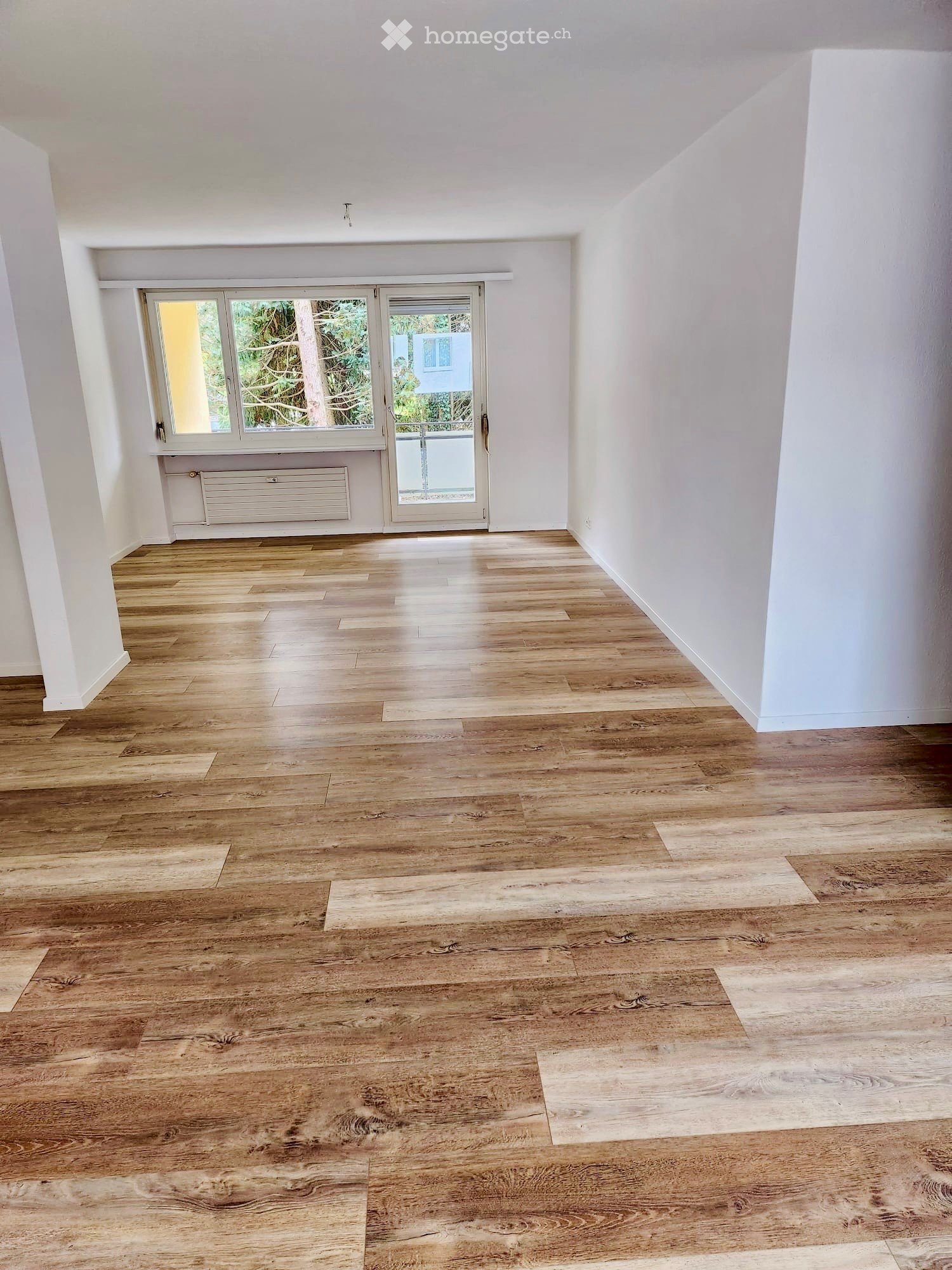 Empty room with wooden flooring, white walls, large window, and sliding glass door leading outside.