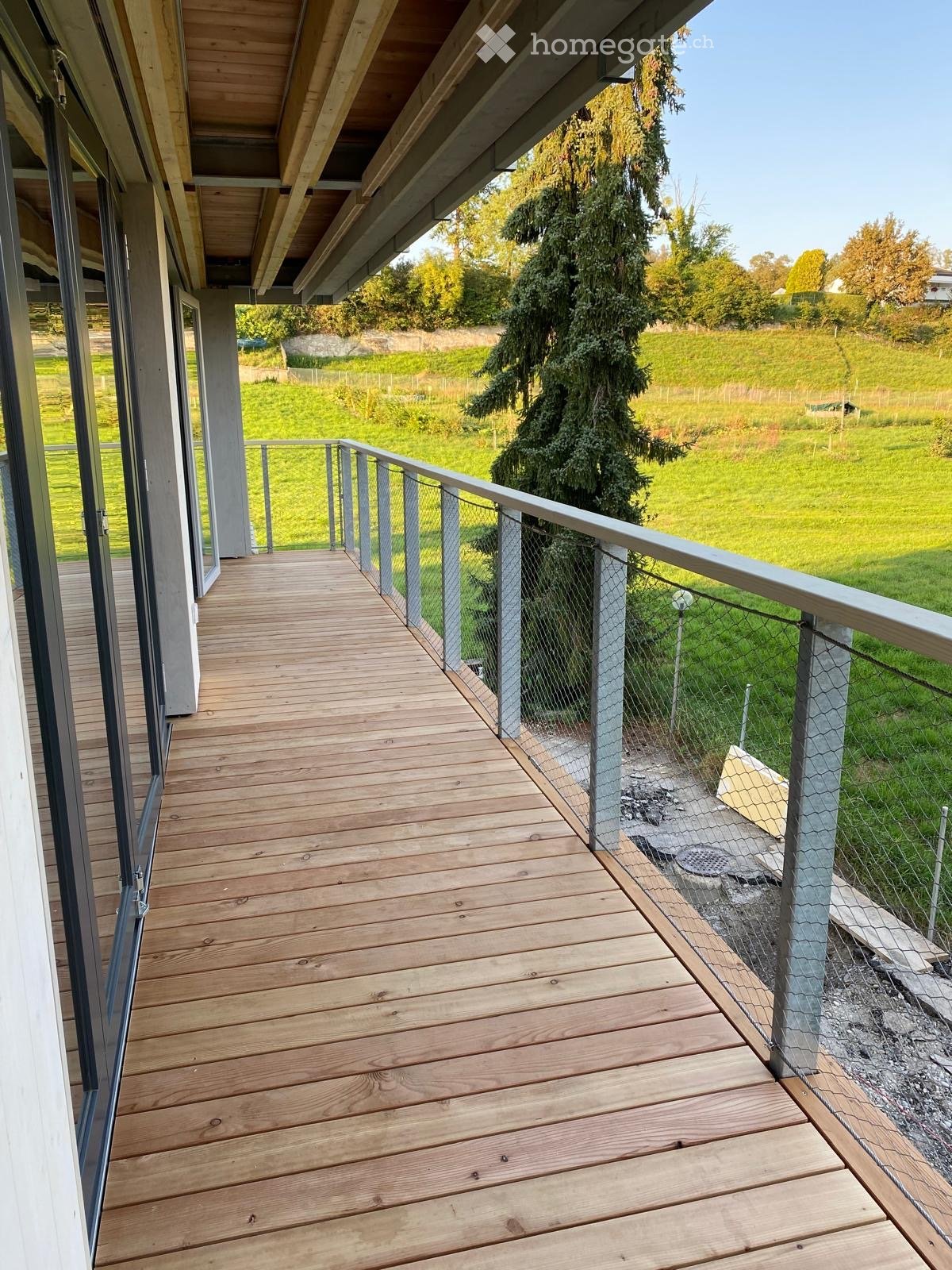 Wooden deck with metal railing, overlooking a grassy area with trees and a fence.