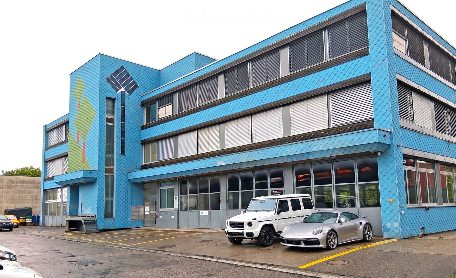 Modern, multi-story building with a bright blue facade and large windows. The building features a contemporary design with solar panels on the roof and a mural on one side. Several luxury cars are parked in front of the building.