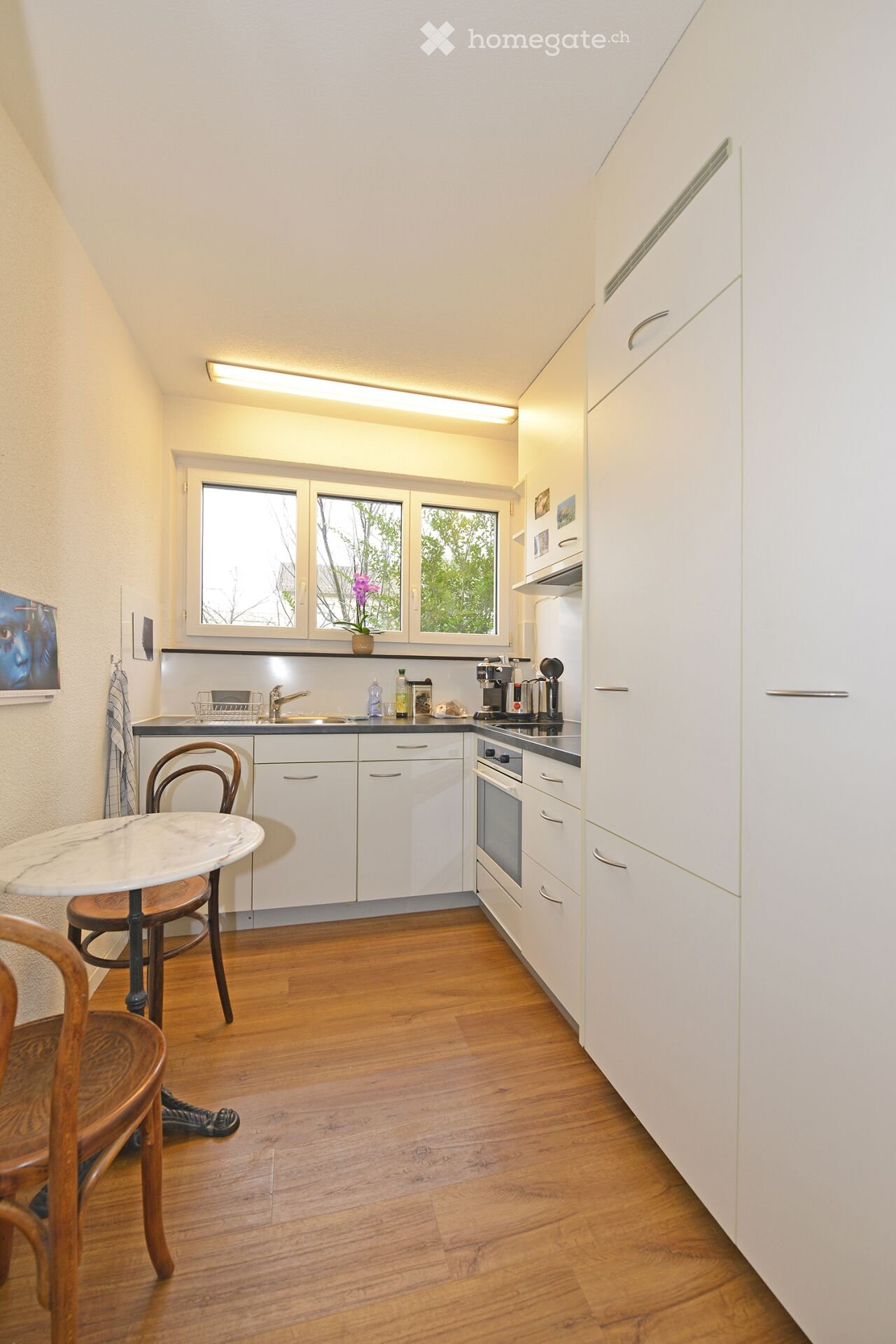 Modern kitchen with white cabinets, wooden flooring, and a small dining area. Features include a sink, oven, coffee maker, and a small table with chairs.