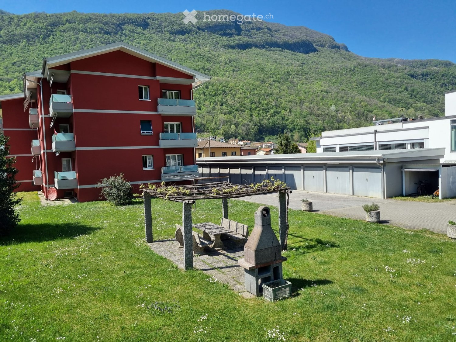Red building with balconies, green lawn, pergola with seating, barbecue, mountain view
