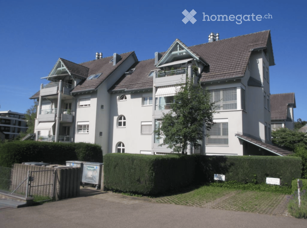 A multi-story residential building with white walls, brown tiled roofs, multiple balconies, and a well-maintained hedge in front.