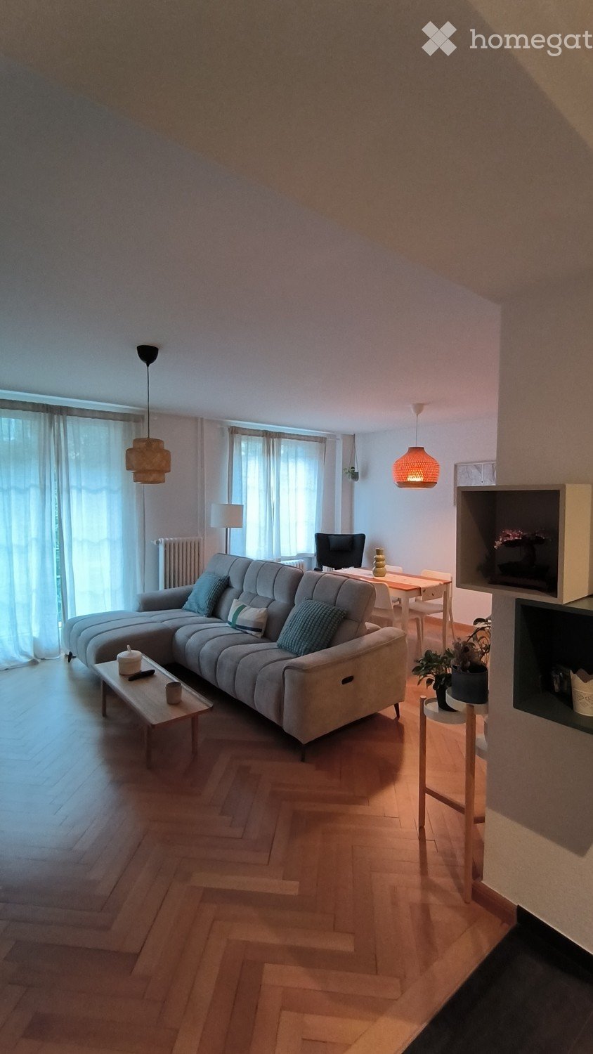 Modern living room with a grey sectional sofa, wooden coffee table, dining area with table and chairs, and a computer on a side table. The room features wooden flooring, large windows with curtains, and pendant lighting.