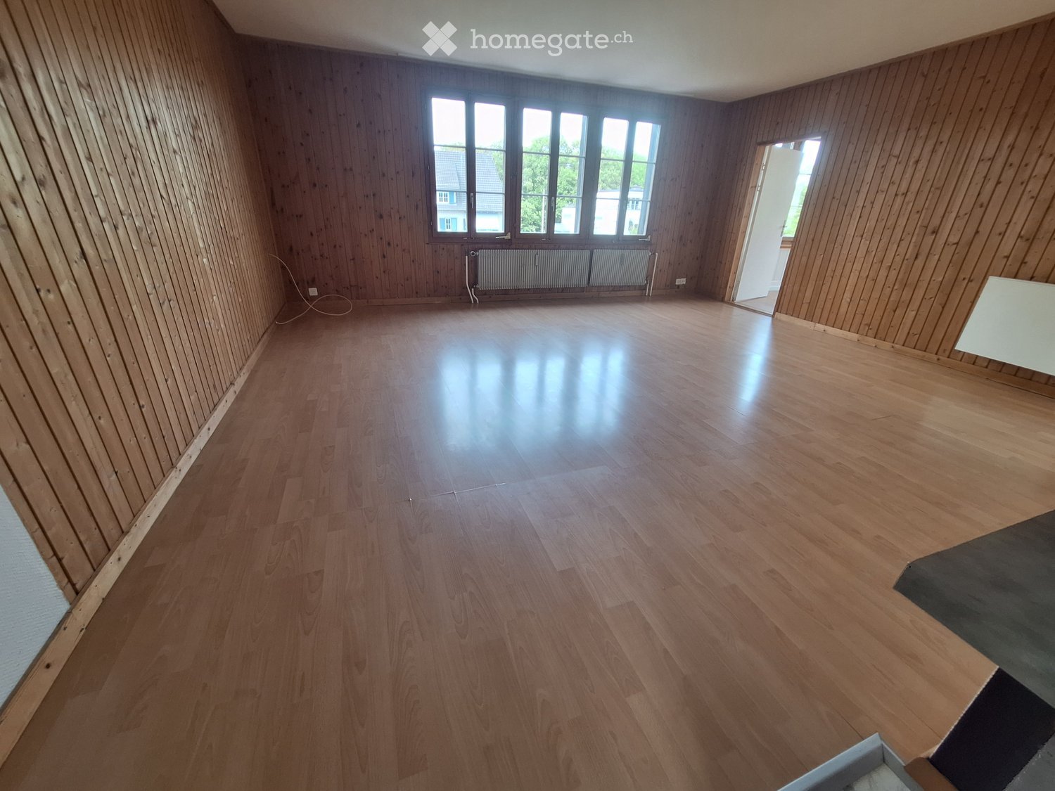 Empty room with wooden walls and floor, large windows, radiator, and an open doorway leading to another space.