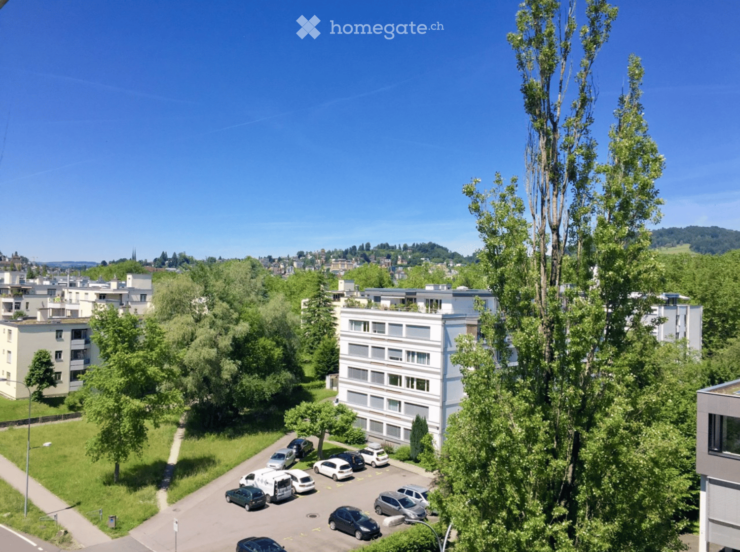 Aerial view of a residential area with multiple apartment buildings, a parking lot with several cars, and surrounding greenery.