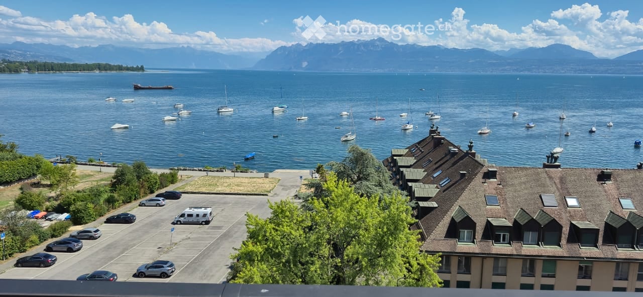 A scenic view of a lake with sailboats, surrounded by mountains, with a parking lot and buildings in the foreground.