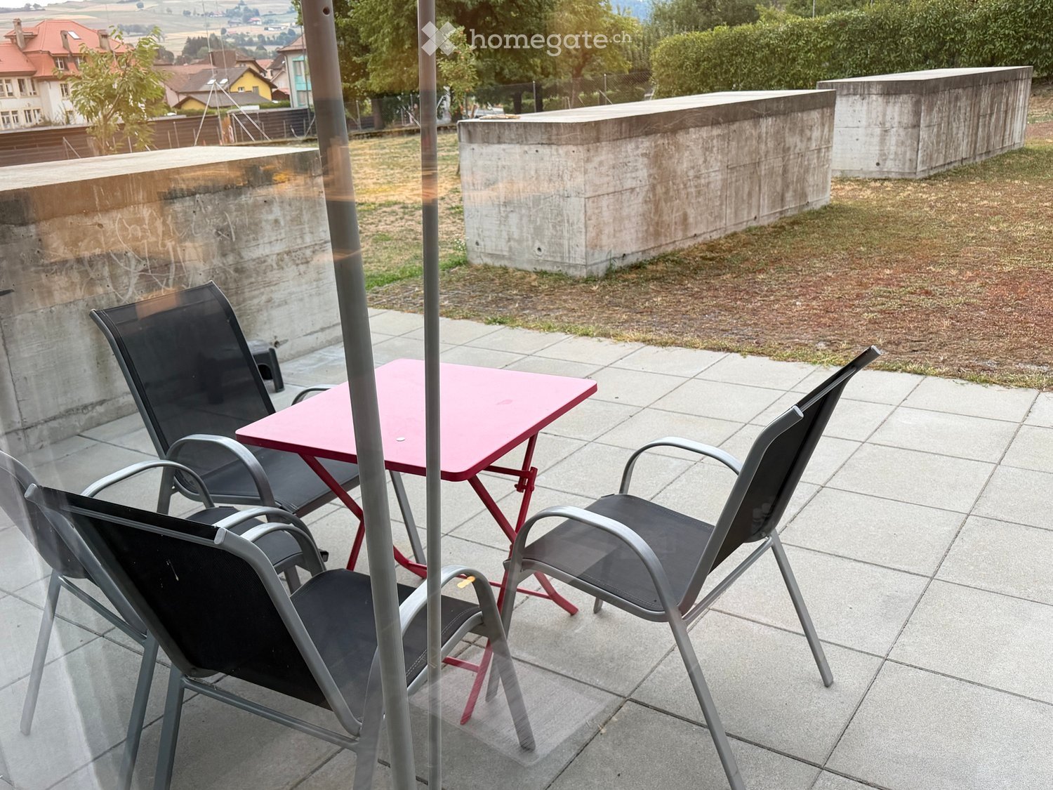 Balcony with tiled floor, pink table, and four black chairs arranged around it. Concrete walls and a grassy area are visible in the background.
