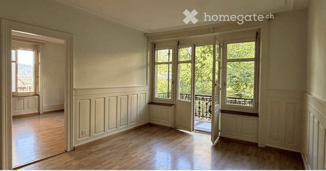 Empty room with wooden flooring, large windows, and a balcony access. Walls are painted in a light color with decorative paneling.