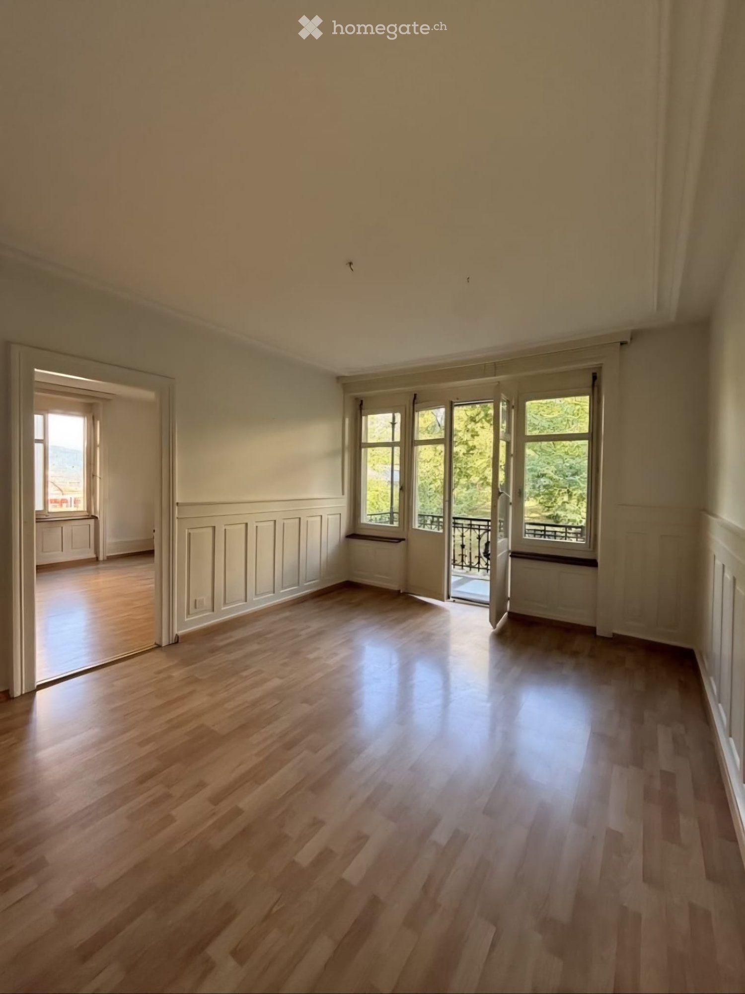 Empty room with wooden flooring, large windows, and a balcony visible through an open door.