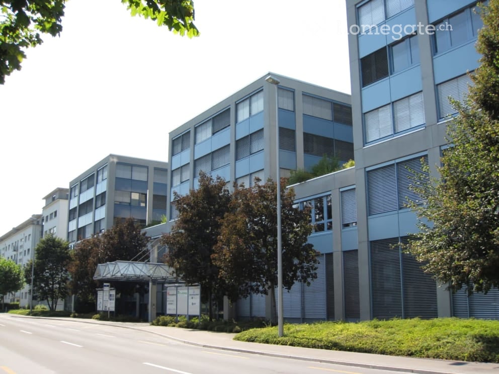 Modern multi-story building with large windows and a covered walkway entrance, surrounded by trees and greenery.
