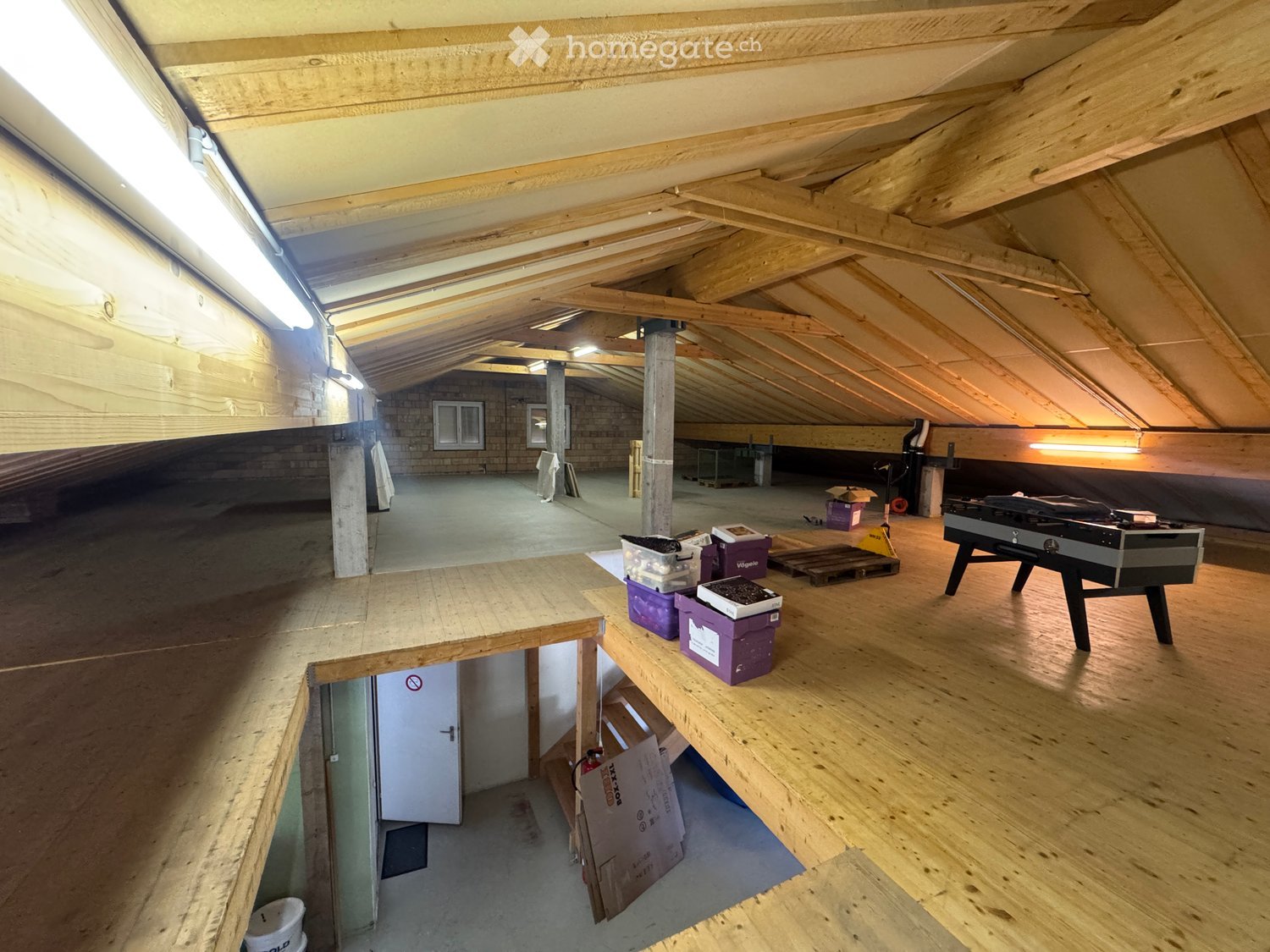 An attic space with wooden beams and exposed rafters, featuring a large wooden table, storage boxes, and a foosball table.