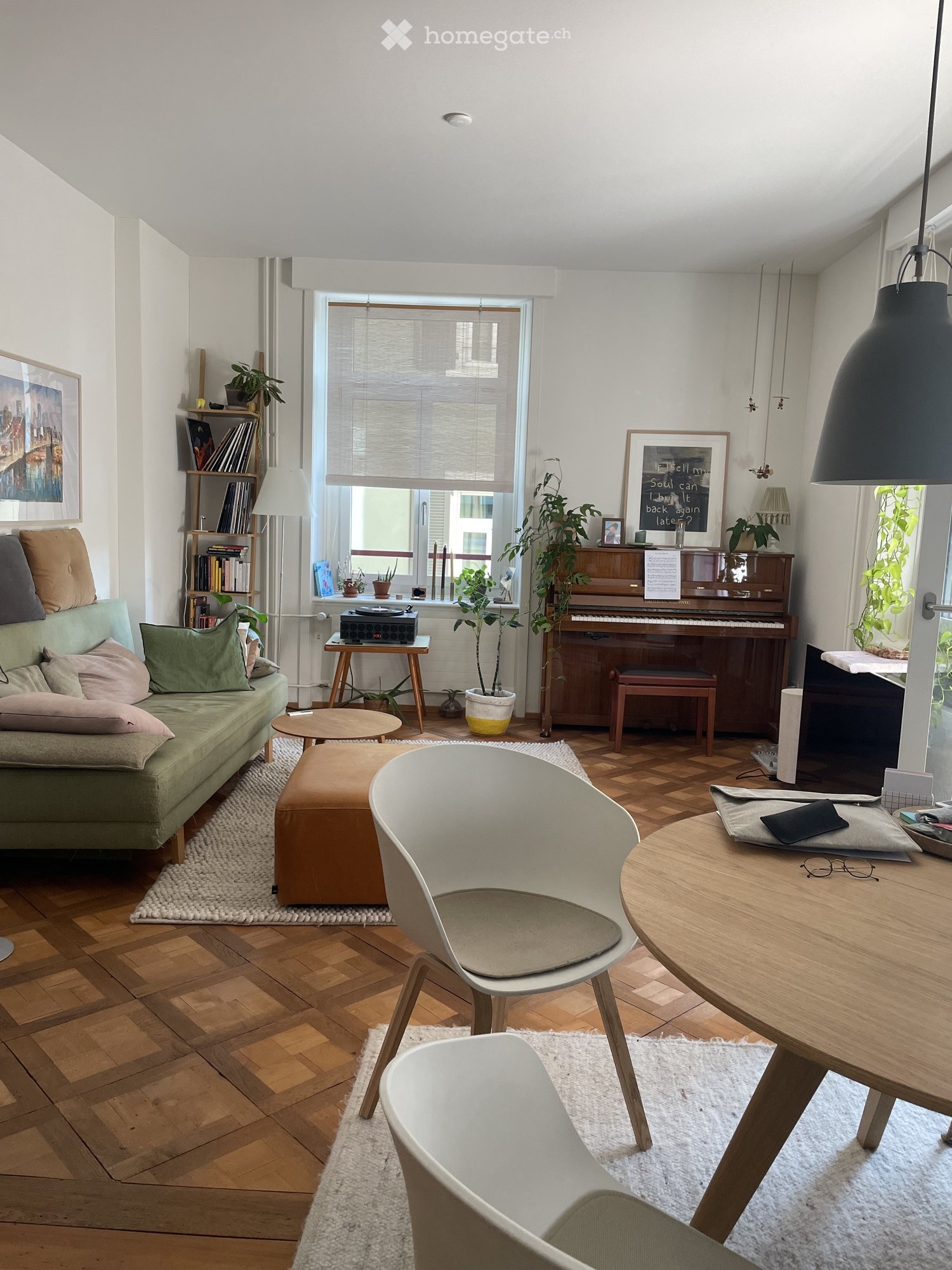 Living room with wooden flooring, large window, green sofa, ottoman, white chairs, round table, piano, bookshelf, and decorative plants.