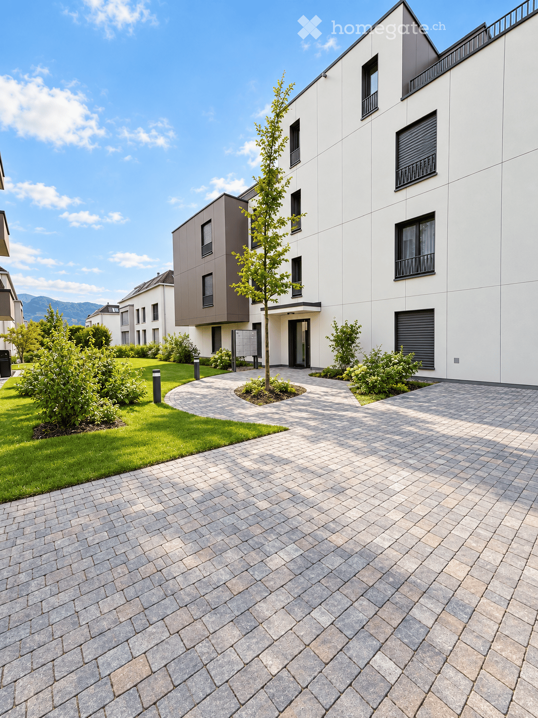 Modern residential building with white and grey facade, multiple windows, and a landscaped entrance area with a paved pathway and small garden.