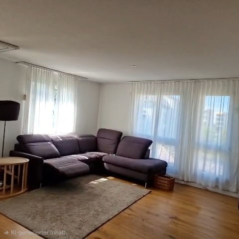 Modern living room with a dark grey sectional sofa, a small round wooden coffee table, a floor lamp, and a large area rug on a wooden floor. Large windows with white curtains allow natural light to fill the space.