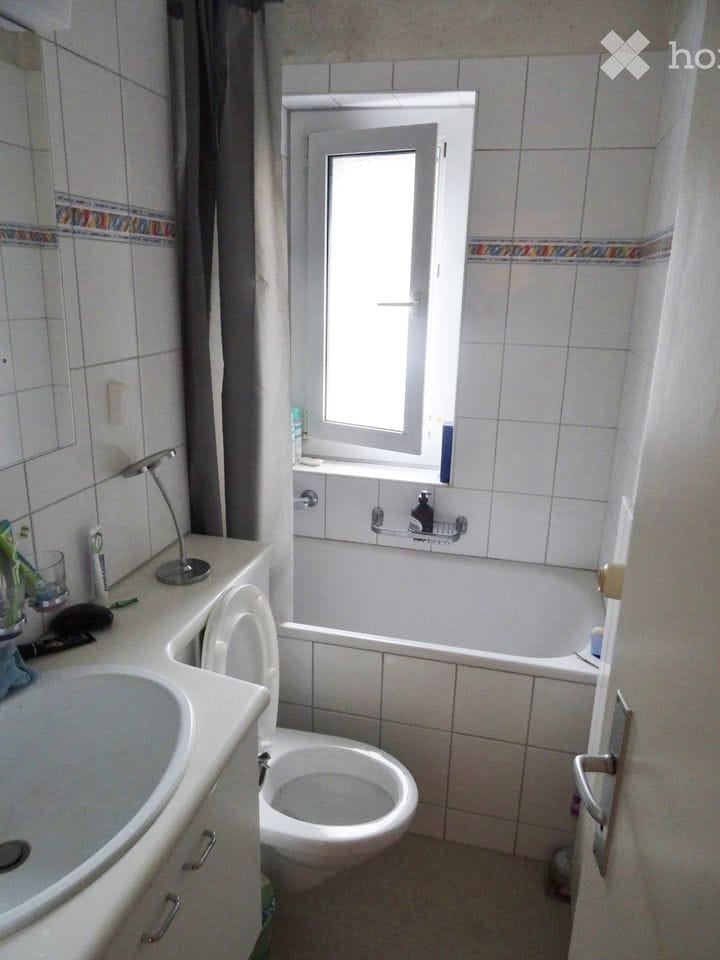 A small bathroom with a toilet, sink, bathtub, and a window. The walls are tiled, and there is a mirror above the sink.