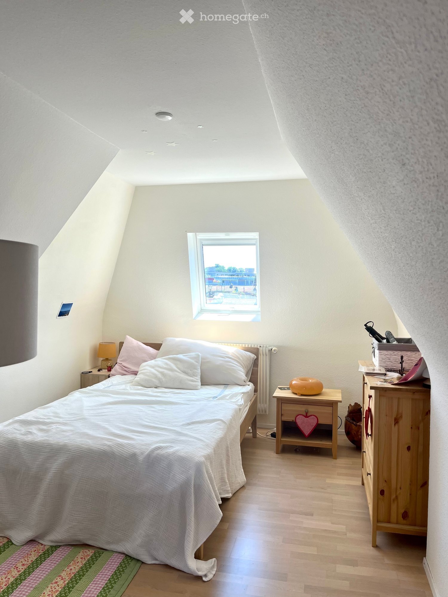Cozy attic bedroom with a single bed, nightstand, lamp, and wooden furniture. Features a skylight window and a small rug on the floor.