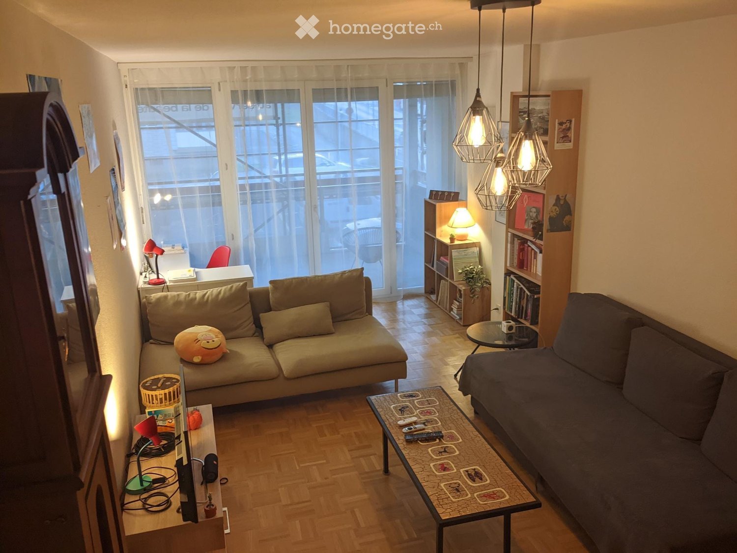 A cozy living room with a sofa, a coffee table, a bookshelf, and a small desk area. The room features wooden flooring, large windows with sheer curtains, and modern pendant lights.